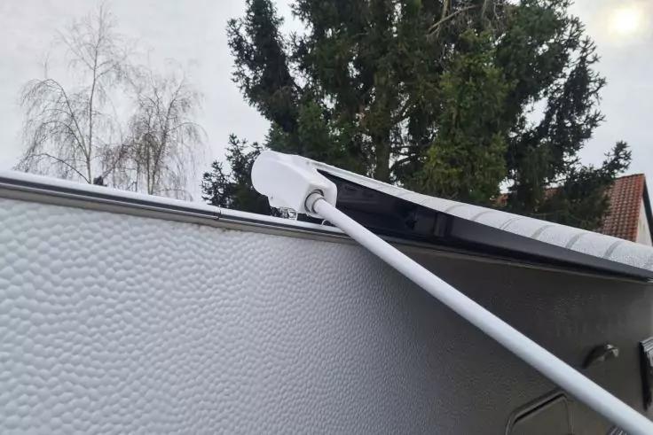 Wei&szlig;er Ausleger einer Markise &uuml;ber der strukturierten silbernen Seitenwand eines Fahrzeugs; im Hintergrund B&auml;ume und ein Hausdach