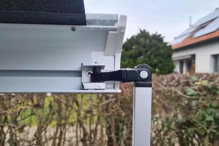 Nahaufnahme eines wei&szlig;en Metallprofils mit schwarzem Gelenk, befestigt an einem senkrechten Metallpfosten; im Hintergrund Hecke und H&auml;user