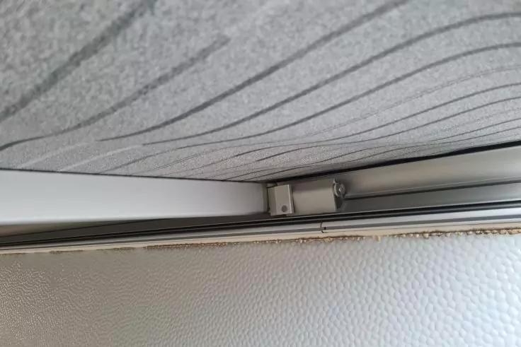 Decken- und Wandkante mit einer Metallf&uuml;hrungsschiene und Befestigungswinkel, dar&uuml;ber hellgraue Deckenverkleidung mit welligem Muster, darunter wei&szlig;e, genarbte Fl&auml;che