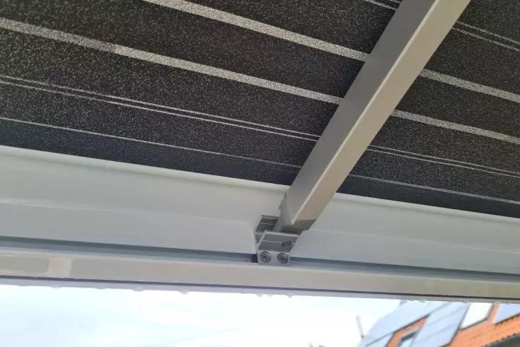 Untere Ansicht eines dunklen Paneels mit parallelen hellen Streifen, wei&szlig;er Metalltr&auml;ger mit Befestigungswinkel und Schrauben, im unteren Bildrand Himmel und Hausd&auml;cher