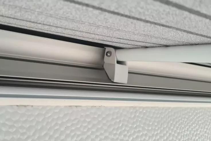 Nahaufnahme einer grauen Metallhalterung mit Schraube an einer oberen Schiene; dar&uuml;ber eine helle, gerillte Fl&auml;che, darunter eine strukturierte wei&szlig;e Fl&auml;che