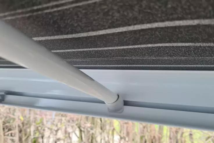 Unterseite eines gestreiften Stoffes, wei&szlig;e Metallstange an einem hellgrauen Rahmen, unscharfer Hintergrund mit Pflanzen