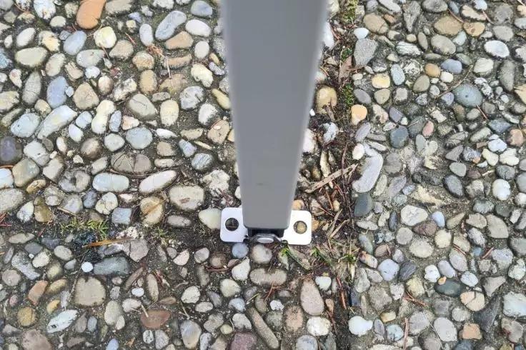 Vertikaler Metallpfosten mit kleiner quadratischer Bodenplatte und zwei sichtbaren Schrauben auf kiesigem Untergrund mit vereinzelten trockenen Bl&auml;ttern