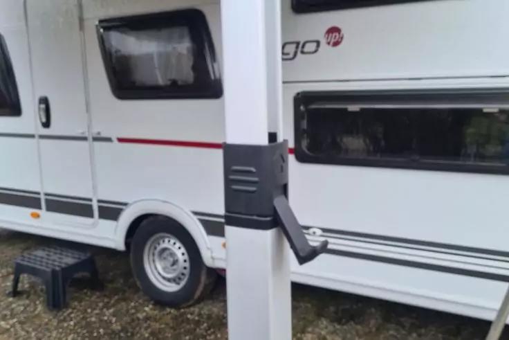 Seitenansicht eines wei&szlig;en Wohnwagens mit Fenstern und Rad; im Vordergrund ein wei&szlig;er Pfosten mit schwarzem Halter, schwarzer Tritt auf Kies