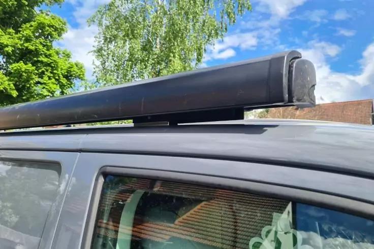 Schwarzes zylindrisches Geh&auml;use auf dem Dachtr&auml;ger eines grauen Autos &uuml;ber der Seitenscheibe; im Hintergrund B&auml;ume und blauer Himmel mit Wolken