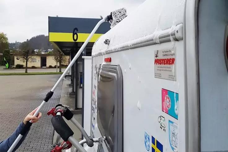 Weiße Fahrzeugseite mit geschlossener Klappe und mehreren Aufklebern, Fahrradträger mit teilweise sichtbarem Lenker und Hand, langer Stab nach oben gerichtet, im Hintergrund Zapfsäule mit gelbem Streifen und Ziffer 6