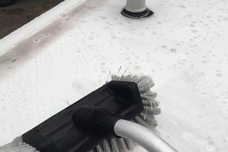 Schwarzer Bürstenkopf mit weißen Borsten an silbernem Griff auf weißer, seifiger Fläche mit Schaum und Wassertropfen; oben rechts rundes Rohr mit schwarzem Dichtungsring