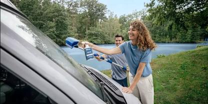 Zwei Personen reinigen die Windschutzscheibe eines wei&szlig;en Fahrzeugs mit einem Abzieher und einem Eimer, im Hintergrund B&auml;ume und Rasen
