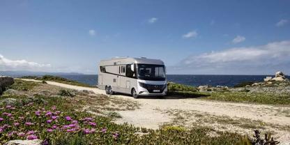 Wei&szlig;es Wohnmobil auf einem unbefestigten Weg an einer felsigen K&uuml;ste; violette Wildblumen im Vordergrund, Meer und blauer Himmel mit wenigen Wolken im Hintergrund