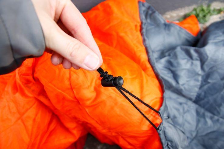 Hand h&auml;lt schwarzen Kordelstopper mit zwei Kordeln vor orange-grauem gestepptem Stoff