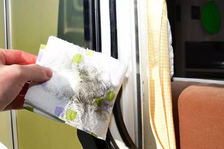 Hand h&auml;lt ein wei&szlig;es Papiertuch mit dunklen Schmutzflecken vor einem Fensterrahmen; gelb kariertes Vorhang und brauner Polstersitz sichtbar