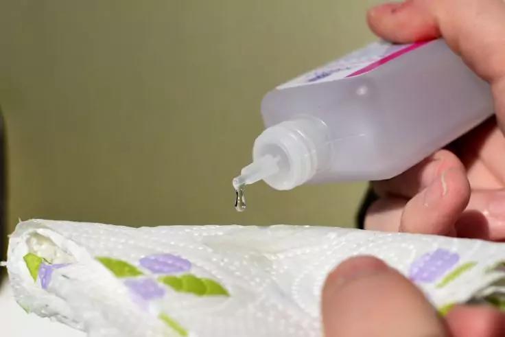 Zwei H&auml;nde halten eine Kunststoffflasche mit T&uuml;lle; ein klarer Tropfen f&auml;llt auf ein gemustertes Papiertuch