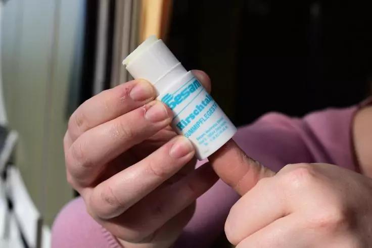 Zwei H&auml;nde halten eine kleine wei&szlig;e Kunststoffdose mit hellblauem Etikett; eine Hand entfernt die Kappe; Person tr&auml;gt rosa &Auml;rmel; verschwommener Hintergrund