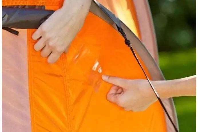 Zwei H&auml;nde an der Au&szlig;enwand eines orangefarbenen Zeltes, eine Hand am oberen Rand, eine andere an einer gen&auml;hten &Ouml;ffnung; schwarzes Spannseil sichtbar, unscharfer gr&uuml;ner Hintergrund