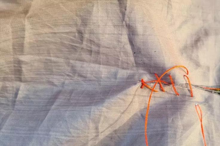 Nahaufnahme eines hellen Stoffs mit orangefarbenem N&auml;hgarn und Nadel, wenige Stiche und lose Fadenenden