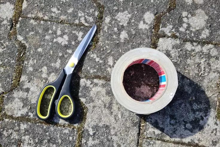 Schere mit schwarzen Griffen und gelben Innenseiten links, Klebebandrolle mit wei&szlig;em Band und buntem Innenring rechts auf grauem, fleckigem Pflaster