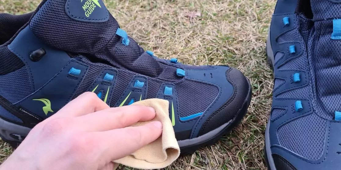Hand wischt einen dunkelblauen Wanderschuh mit einem beigen Tuch, ein zweiter Schuh steht daneben auf Gras
