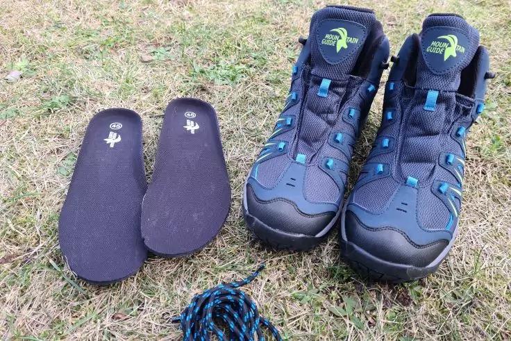 Zwei blau-schwarze Wanderschuhe rechts, zwei schwarze Einlegesohlen links, zusammengerollte blau-schwarze Schn&uuml;rsenkel vorne, alles auf trockenem Gras