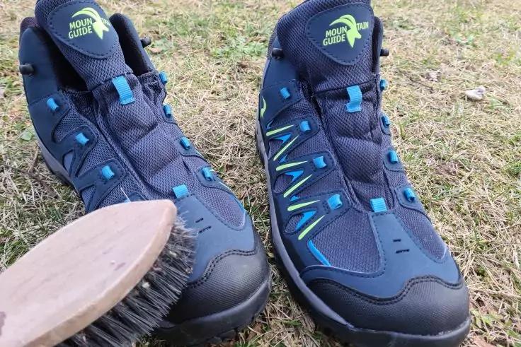 Zwei dunkle Wanderschuhe mit blauen Akzenten auf Gras; vorne links liegt eine Schuhb&uuml;rste mit Holzgriff