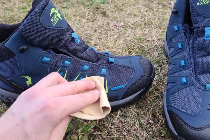 Hand mit gelbem Tuch an der Vorderseite eines blauen Wanderschuhs; ein zweiter blauer Schuh steht daneben auf Gras