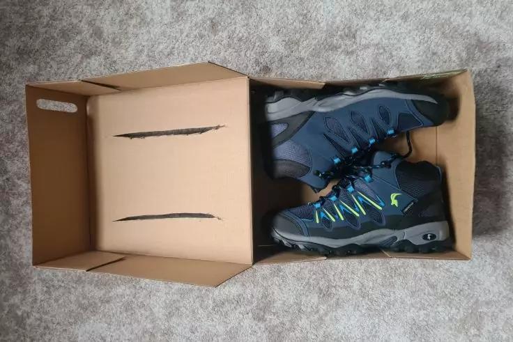 Offener brauner Karton auf grauem Teppich, darin ein Paar dunkelblaue Schuhe mit hellblauen und neongr&uuml;nen Details