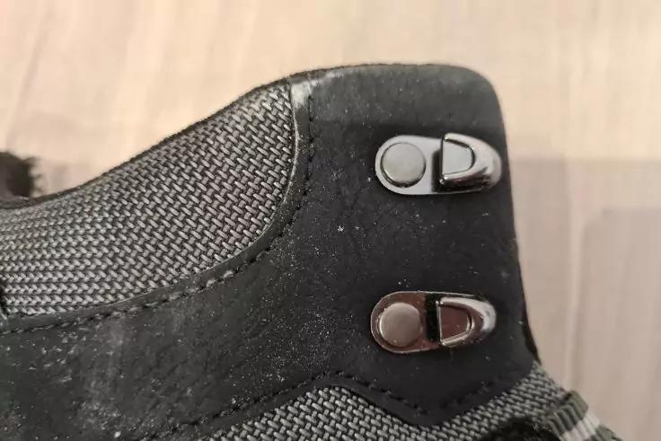 Nahaufnahme eines schwarz-grauen Schuhs mit zwei metallischen Haken&ouml;sen, sichtbare Stoffstruktur, heller Holzboden im Hintergrund