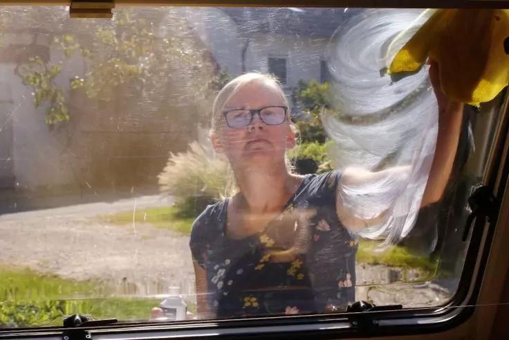 Person mit kurzen hellen Haaren und Brille wischt die Au&szlig;enseite eines Fensters mit einem gelben Tuch; im Hintergrund Gras, ein Weg und ein Geb&auml;ude