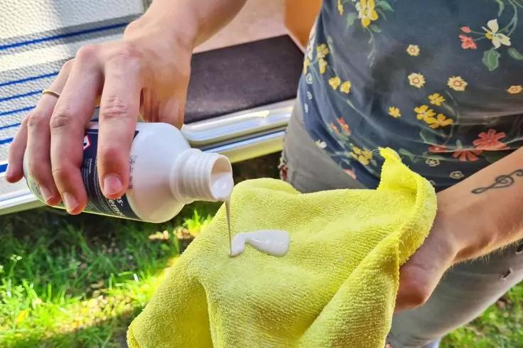 Zwei H&auml;nde dr&uuml;cken eine Flasche, wei&szlig;e Fl&uuml;ssigkeit tropft auf ein gelbes Tuch; gebl&uuml;mtes Oberteil und Tattoo am Unterarm sichtbar, im Hintergrund Gras