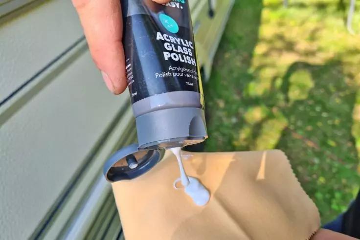 Hand dr&uuml;ckt eine Tube mit der Aufschrift "Acrylic Glass Polish"; wei&szlig;e Paste auf einem beigefarbenen Tuch, Hausfassade und Rasen im Hintergrund