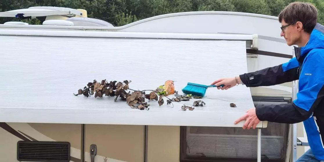 Mann in blauer Jacke fegt Bl&auml;tter und Schmutz von der wei&szlig;en Markise eines Wohnwagens