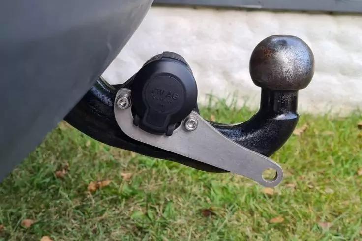 Nahaufnahme einer Anh&auml;ngerkupplung mit Anh&auml;ngerkugel, schwarzer Steckdose mit Schutzkappe und silberner Halterung; Gras und wei&szlig;e Mauer im Hintergrund