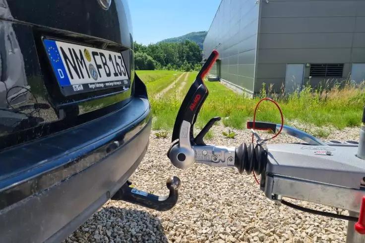 Heck eines schwarzen Autos mit Anh&auml;ngerkupplung, Anh&auml;ngerdeichsel und rotem Handbremsgriff; Kiesboden, Geb&auml;ude und B&auml;ume im Hintergrund