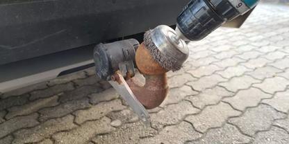 Rostige Anh&auml;ngerkupplungskugel unter dem Heck eines Fahrzeugs, eine Bohrmaschine mit Drahtb&uuml;rstenaufsatz liegt an, gepflasterter Boden darunter