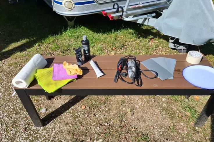 Braune Holzbank mit schwarzem Gestell im Freien. Auf der Bank: K&uuml;chenpapierrolle, gr&uuml;nes und violettes Tuch, gelbes Tuch, schwarze Sprayflasche, grau-schwarzes Elektrowerkzeug mit Kabel, mehrere graue Bl&auml;tter, wei&szlig;e Schale und blauer Teller; im Hinte