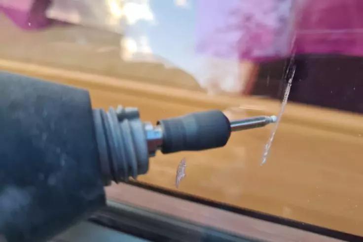 Nahaufnahme eines Handwerkzeugs mit kleinem Bohrer an einer Glasscheibe; zwei senkrechte Risse im Glas; Holzfensterbank und unscharfer lila Reflex im Hintergrund