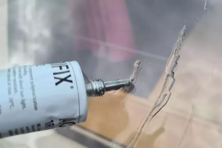 Wei&szlig;er Tubus mit Metalld&uuml;se, der klaren Klebstoff in Linien auf eine transparente Fl&auml;che auftr&auml;gt; unscharfer Hintergrund