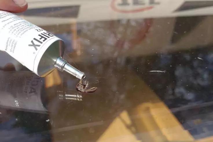 Hand h&auml;lt eine Tube mit Metallspitze; klarer Gel-Tropfen auf gl&auml;nzender Glasfl&auml;che, Spiegelung und unscharfer Hintergrund