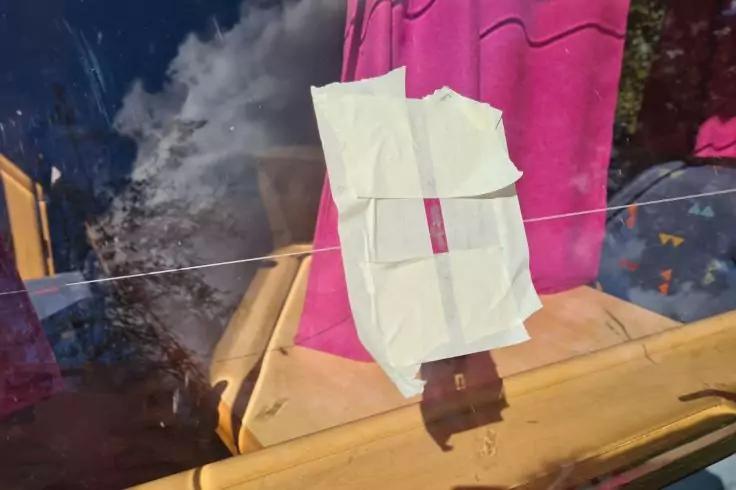 Wei&szlig;es Papier mit Klebeband an einer Autoscheibe, rosa Stoff dahinter, gelber Fahrzeugrahmen und Wolkenreflexion im Glas