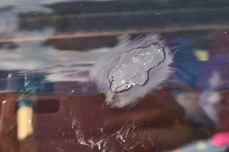 Nahaufnahme einer unregelm&auml;&szlig;igen hellen Stelle auf einer gl&auml;nzenden dunklen Oberfl&auml;che, mit Kratzspuren und unscharfen bunten Spiegelungen im Hintergrund