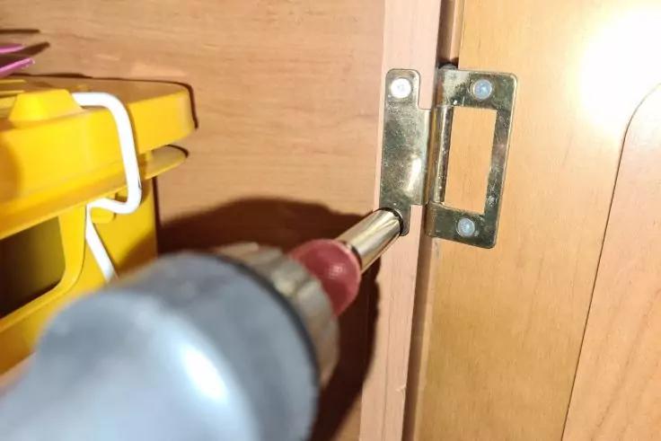 Akkuschrauber-Bit in einer Schraube einer gl&auml;nzenden Metallscharnierplatte an der Kante eines Holzschranks; links ein gelber Kunststoffbeh&auml;lter