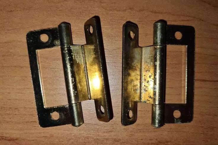 Zwei kleine Messingscharniere auf einer Holzoberfl&auml;che, leicht angelaufen, jeweils zwei rechteckige Platten mit Schraubl&ouml;chern und einem zylindrischen Zapfen