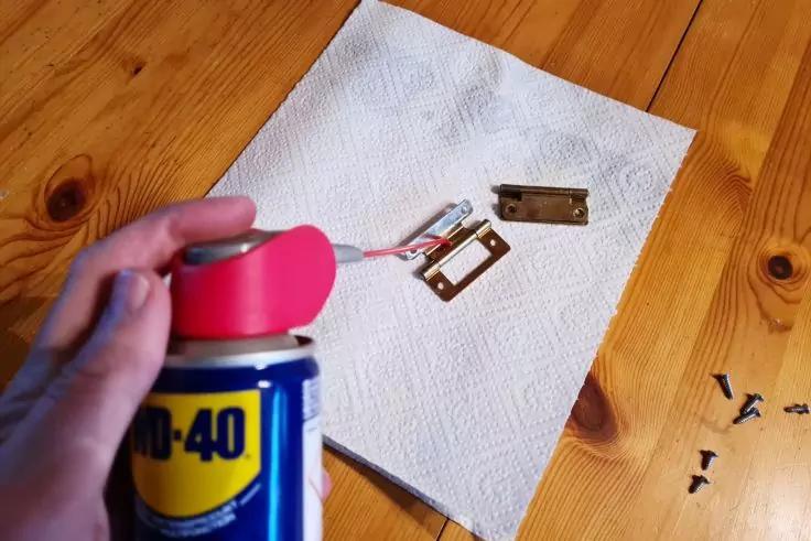 Hand h&auml;lt eine WD-40-Dose mit pinkem Spr&uuml;hrohr &uuml;ber einem Papiertuch, auf dem zwei Metallteile liegen; rechts verstreute kleine Schrauben auf einem Holztisch