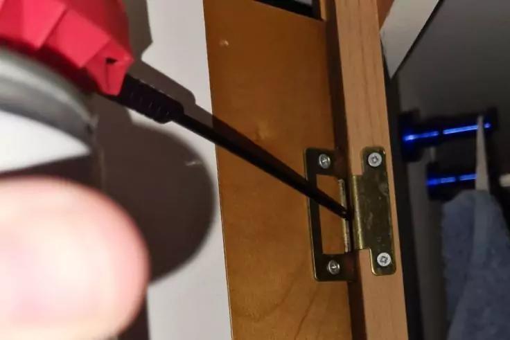 Nahaufnahme: Schraubendreherspitze in einer Schraube eines Metallbands an einer Holzklappe; linker Daumen im Vordergrund; rechts ein Handtuch und ein dunkles M&ouml;belst&uuml;ck