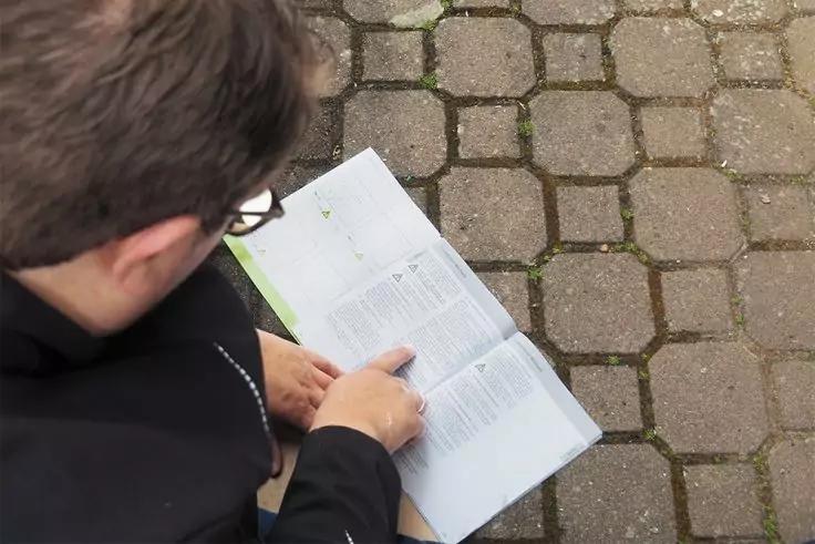 Person von oben mit dunkler Jacke, kurzen Haaren und Brille zeigt mit dem Finger auf ein geöffnetes Buch oder Magazin, das auf gepflastertem Boden liegt