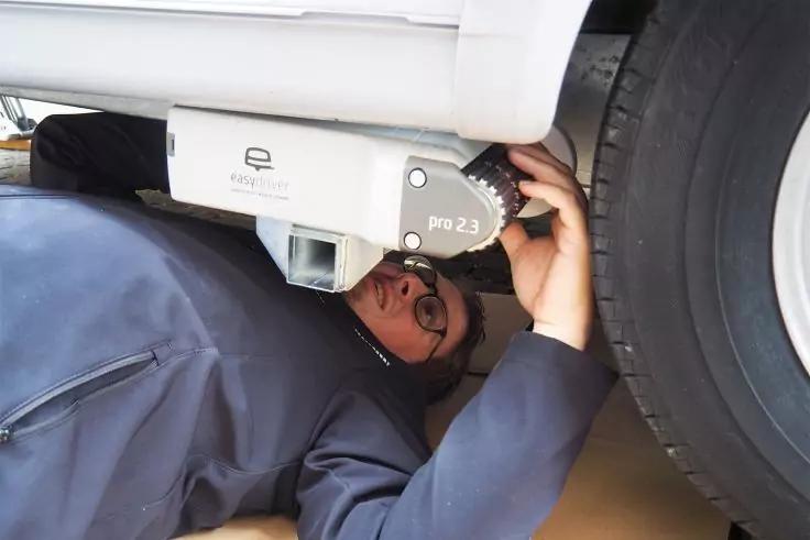 Person liegt auf dem Rücken unter einem Fahrzeug neben einem Rad und hält ein weißes Gerät mit der Aufschrift ‚pro 2.3‘; trägt Brille und dunkle Jacke