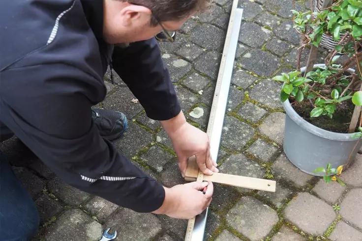 Person kniet auf Kopfsteinpflaster, hält Lineal und Holzstück an einer Metallstange; Topfpflanze rechts