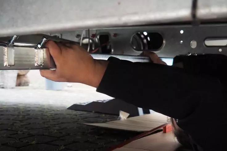 Zwei Hände in dunklen Ärmeln halten ein Metallteil unter der Unterseite eines Fahrzeugs; gepflasterter Boden, Karton und ein Schraubenschlüssel liegen daneben