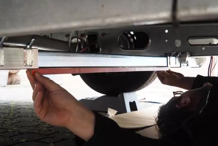 Person unter Fahrzeugrahmen hält mit beiden Händen eine Metallstange; rundes Rad im Hintergrund, Kopf mit Brille rechts, gepflasterter Boden