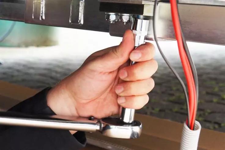 Hand hält Steckschlüssel an Schraube unter Metallrahmen; rote und weiße Kabel daneben