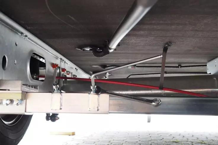 Metallrahmen und Querträger unter einer Plattform, rotes Kabel, mehrere Metallrohre und Befestigungen, links teilweise sichtbares Rad, gepflasterter Boden